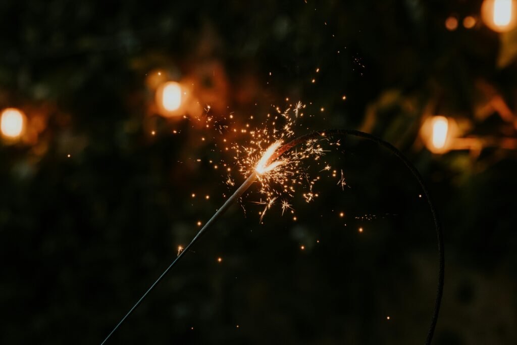 A close-up of a sparkler glowing amid warm, bokeh lights in a dark setting.